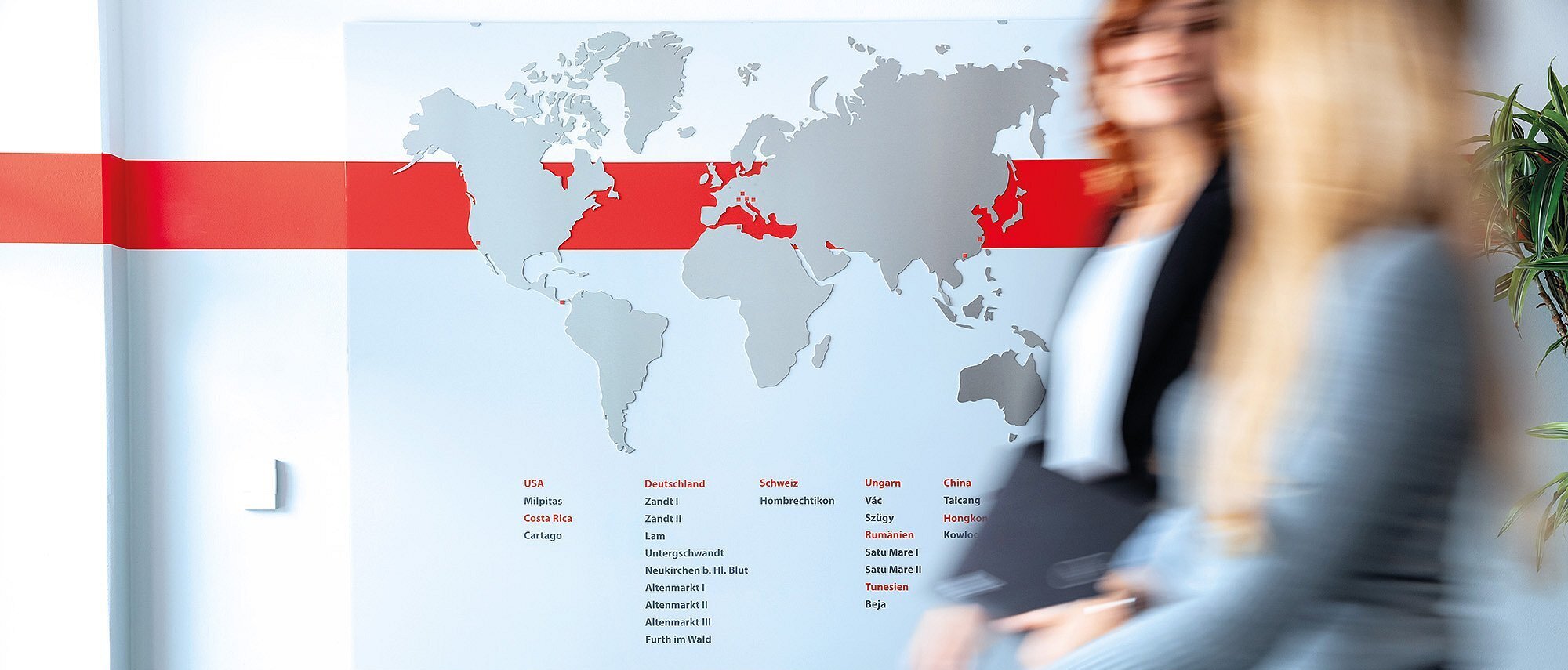 Zwei Mitarbeiterinnen der Zollner Elektronik AG gehen durch einen Flur vor einer Weltkarte im Bereich Global Procurement.